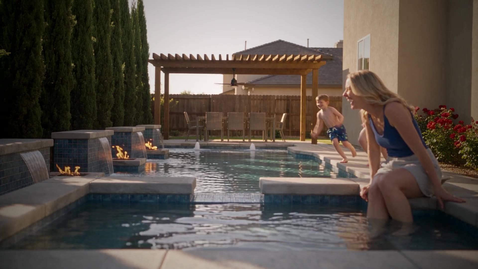 A mom sits on the pool edge with her feet in the water, laughing as her kids jump in behind her. A Story film made from a pool render.