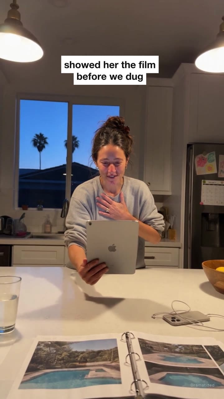 A woman in her kitchen holds a tablet to her chest, smiling in surprise as she watches a video of her backyard pool.